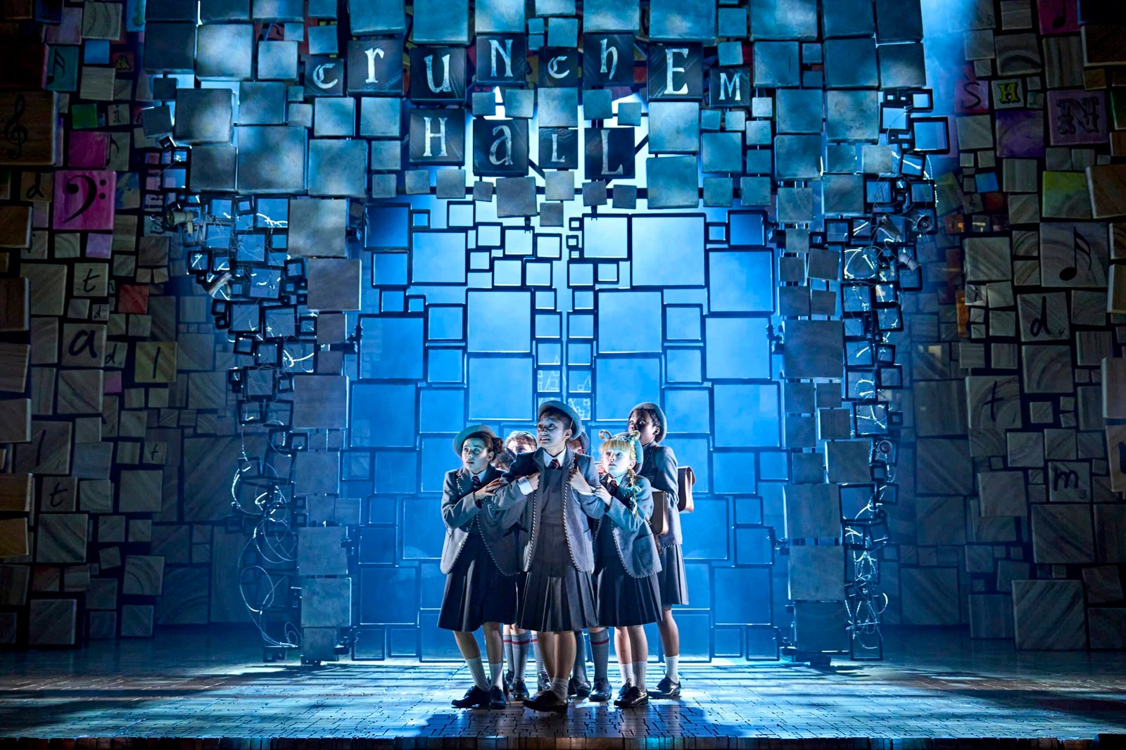 children on stage for matilda the musical. dressed in grey school uniforms and huddled together under blue light