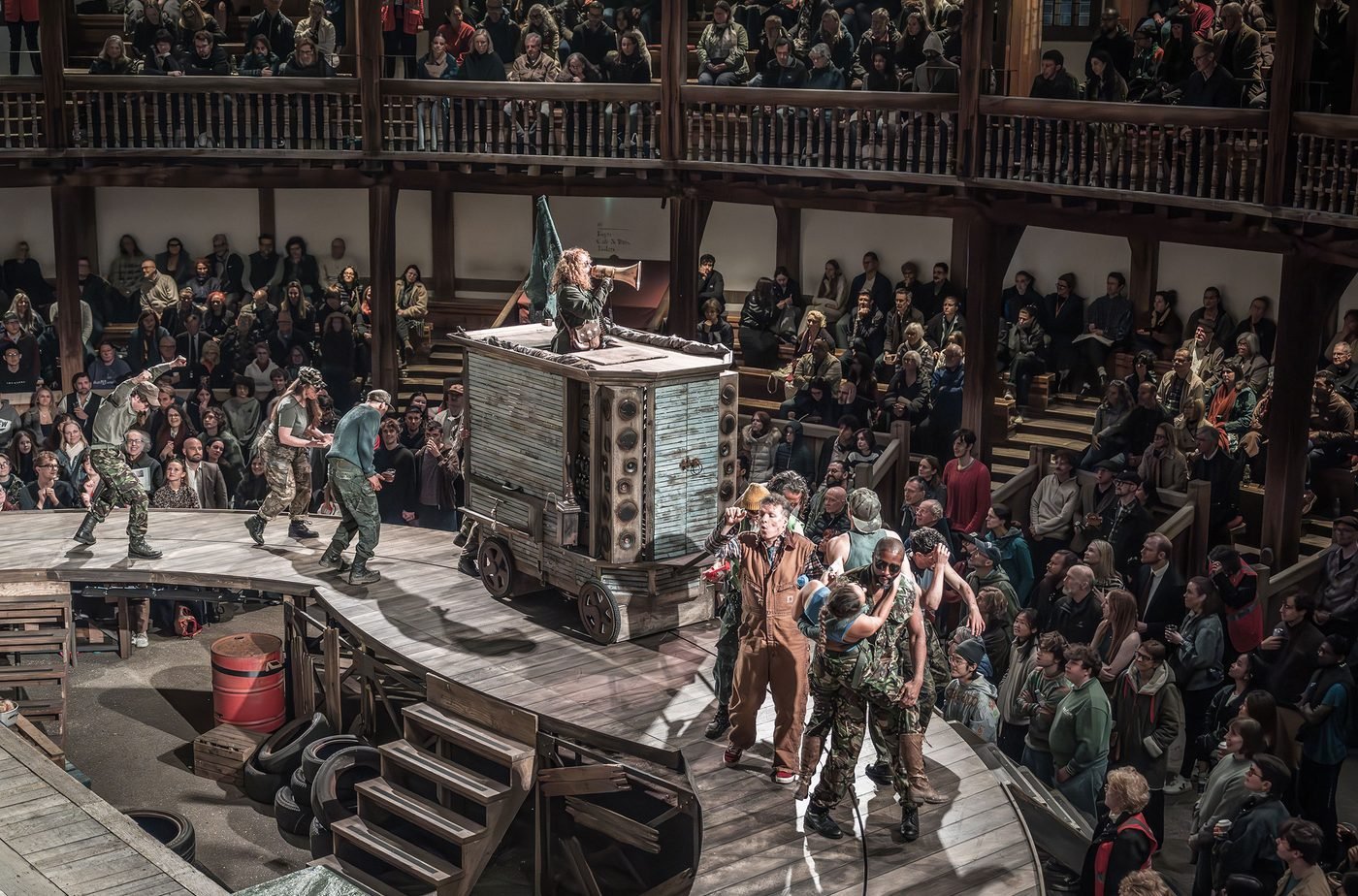 cast members at shakespeare's globe theatre in mother courage and her children. crowds of people with one woman on top a carriage