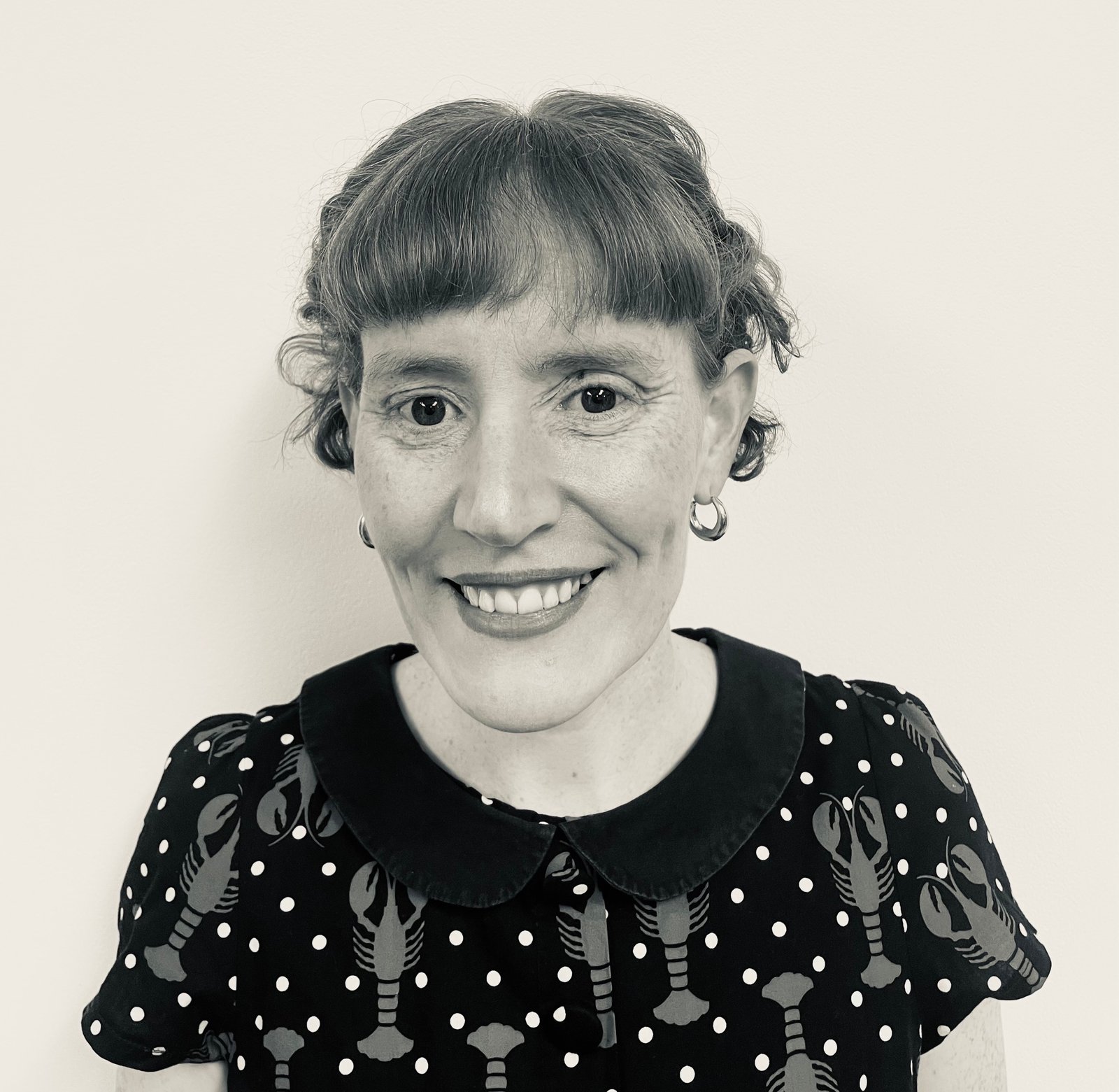 Black-and-white portrait of author Rosa Campbell smiling at the camera while wearing a patterned dress with a dark collar, photographed for The Women's Journal interview for The Book that Taught the World to Orgasm and then Disappeared review