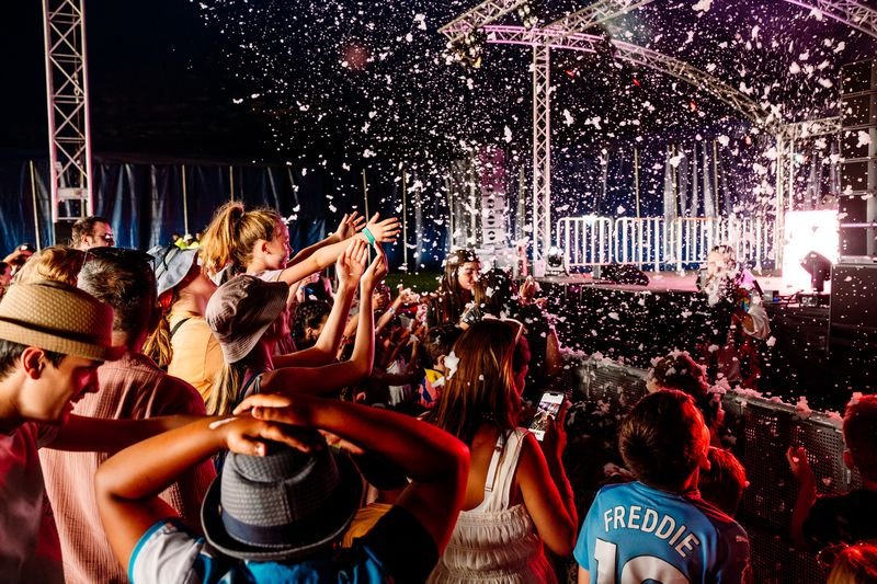 A lively crowd, including children, cheers with upraised hands near a lit stage as fake snow or foam rains down on them during a night performance at the Neighbourhood on Blackheath.