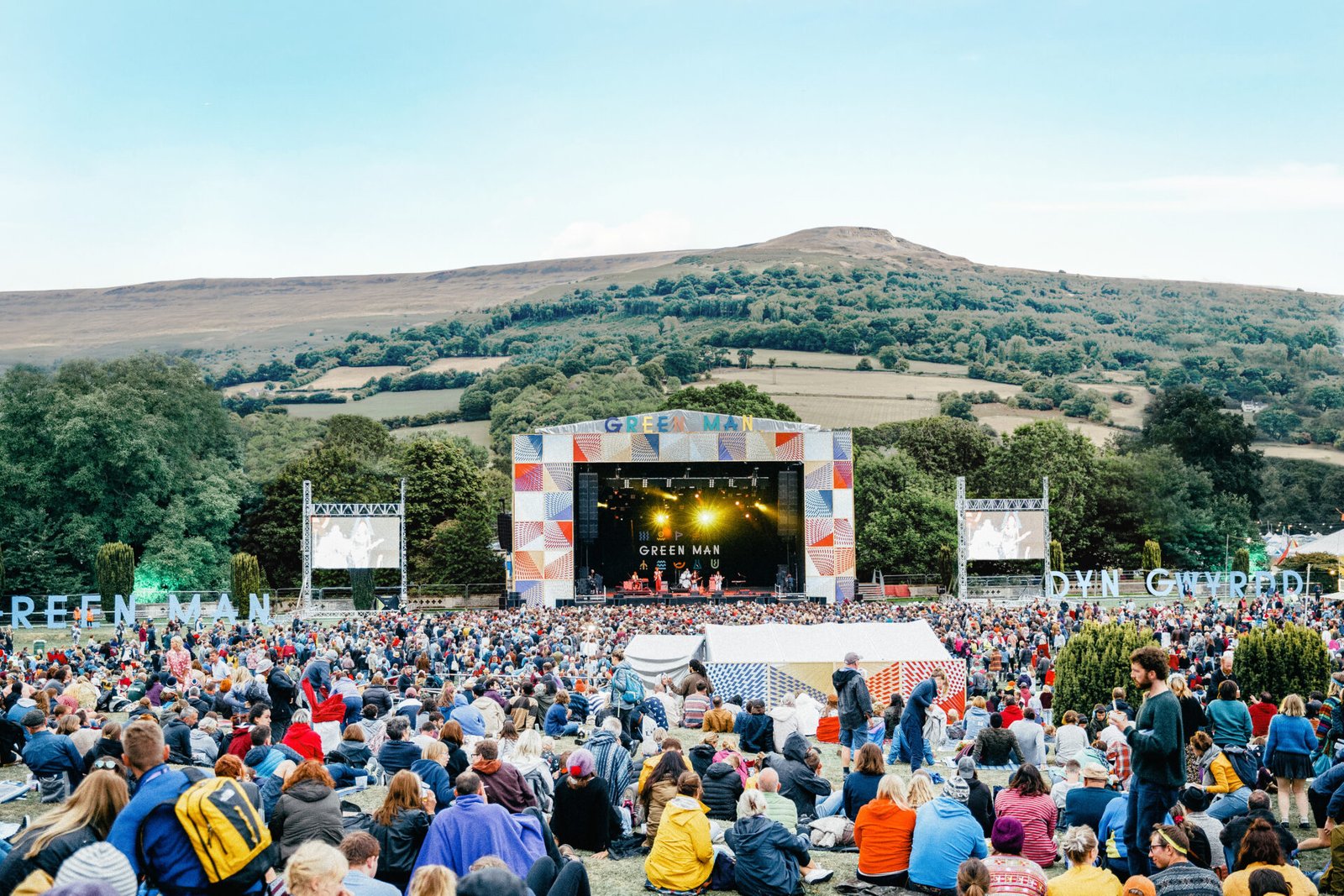 A massive crowd sits on a grassy hill watching a performance on a colorful, large outdoor stage, set against a backdrop of rolling green mountains at the Green Man Festival.