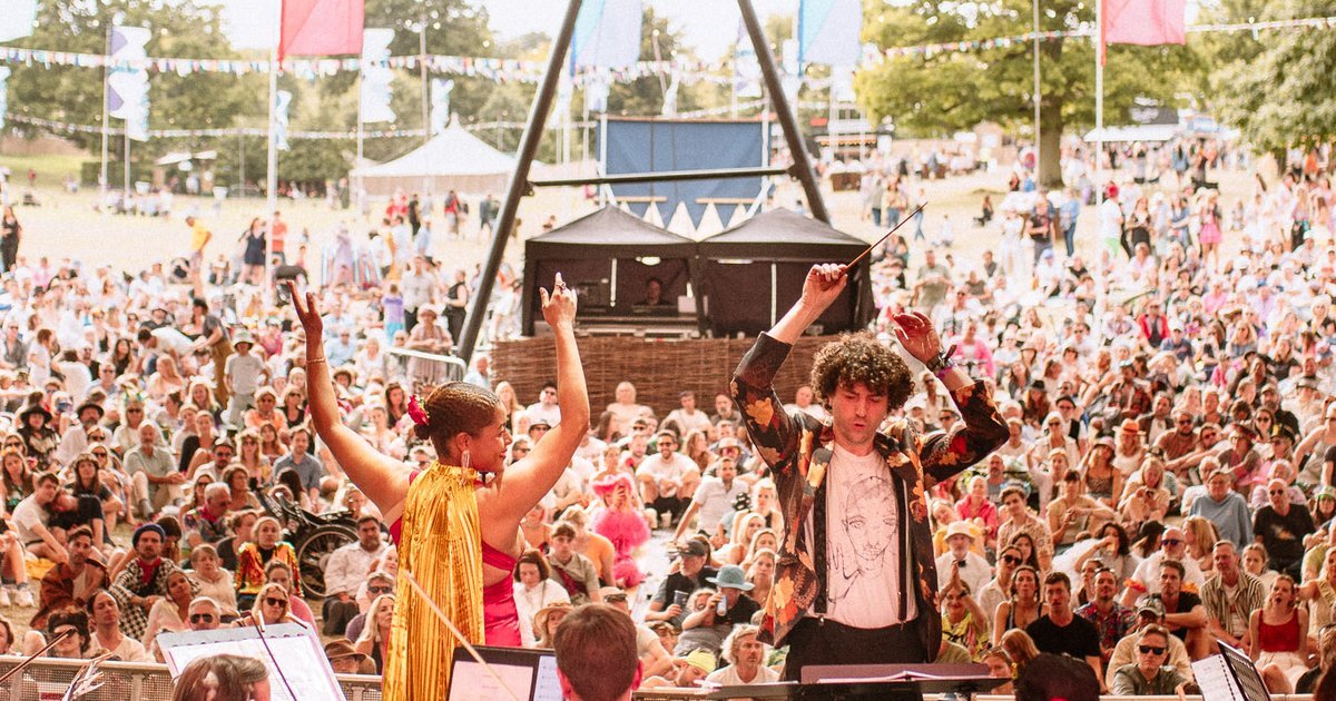 An orchestral conductor and a performer in a gold cape stand on an outdoor stage with their arms raised, leading a performance. In the background, a large, dense crowd of festival-goers sits on a grassy hill under daytime sky at the Wilderness Festival.