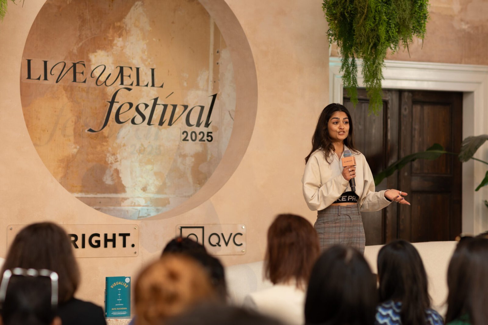 A woman stands on a stage speaking into a microphone to an audience. Behind her is a textured beige wall featuring a large circular "Live Well Festival 2025" logo.