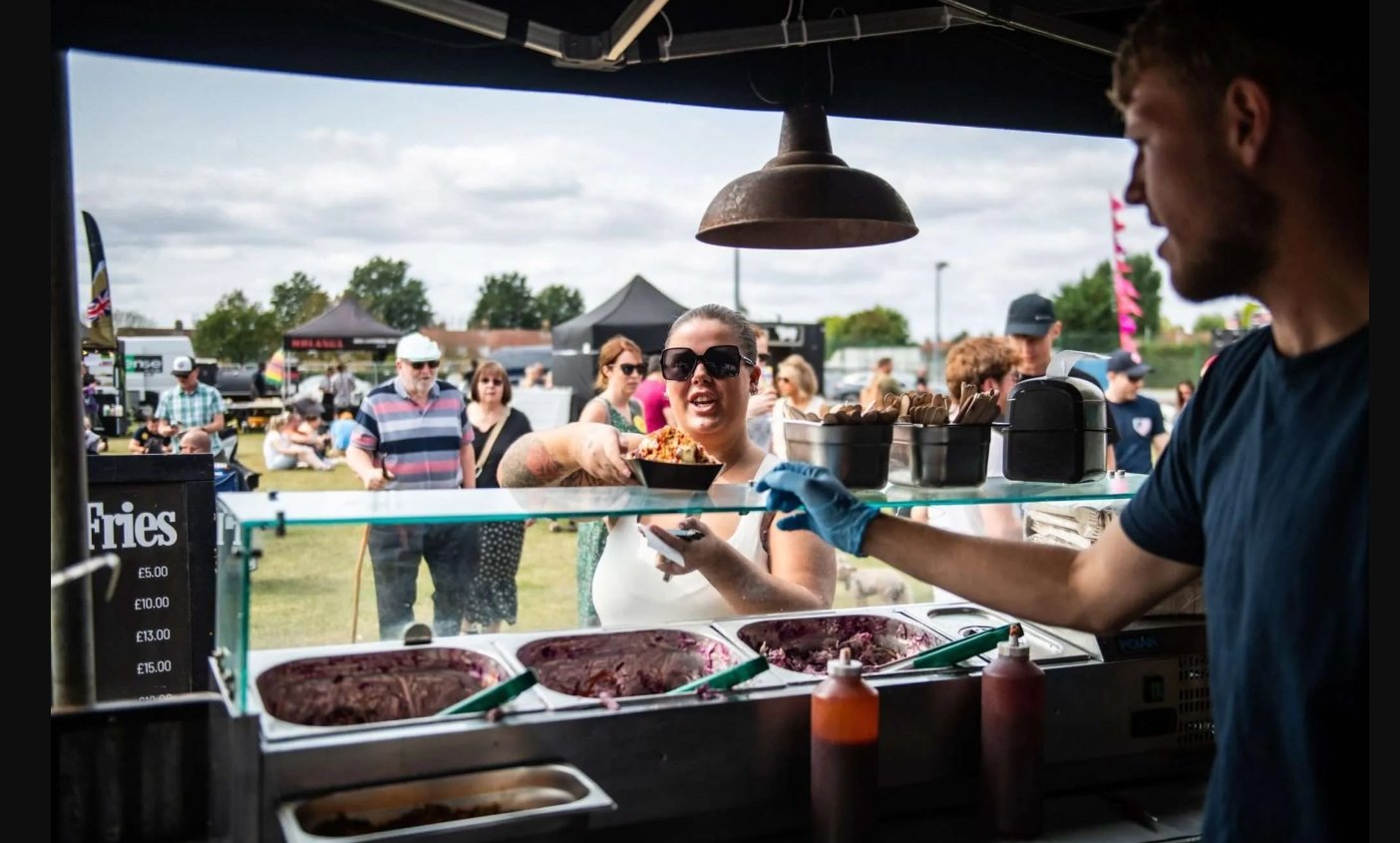 A woman in sunglasses receives a container of food from a vendor under a tent at the Smoke and Fire Festival, with an outdoor festival crowd blurred in the background.
