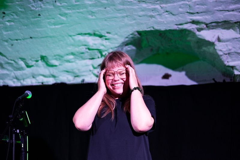 Alfrun Rose smiles with her hands to her head, glasses on, and standing next to a microphone while performing in new London play Dead Air