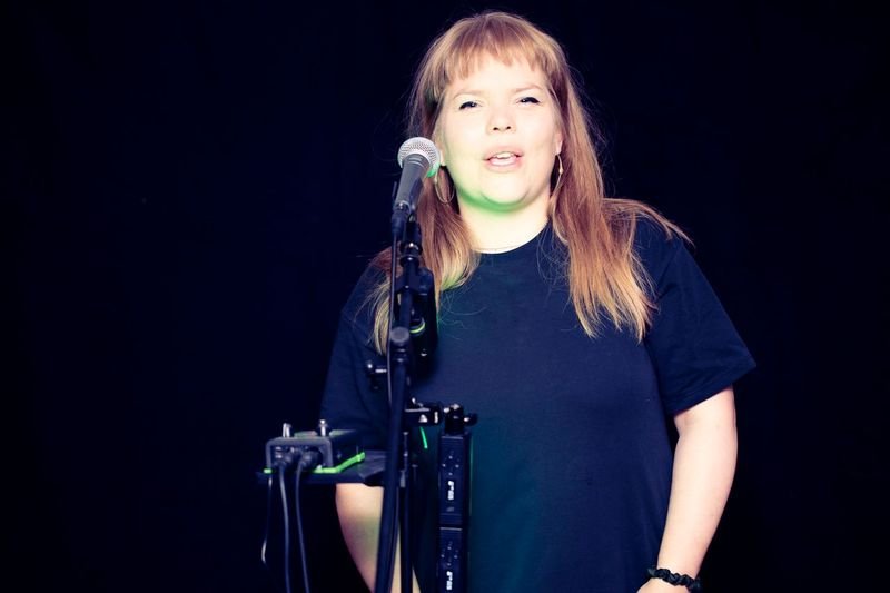 Alfrun Rose speaks into a microphone while performing Dead Air play on a stage with a black background