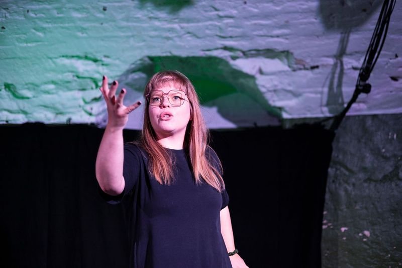 Alfrun Rose performs on stage during Dead Air at Greenwich Theatre, gesturing toward the audience beneath green lighting.