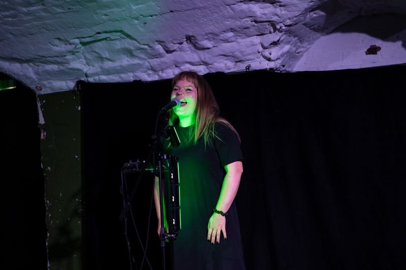 Alfrun Rose speaks enthusiastically into a microphone on a stage lit by green light in Dead Air play