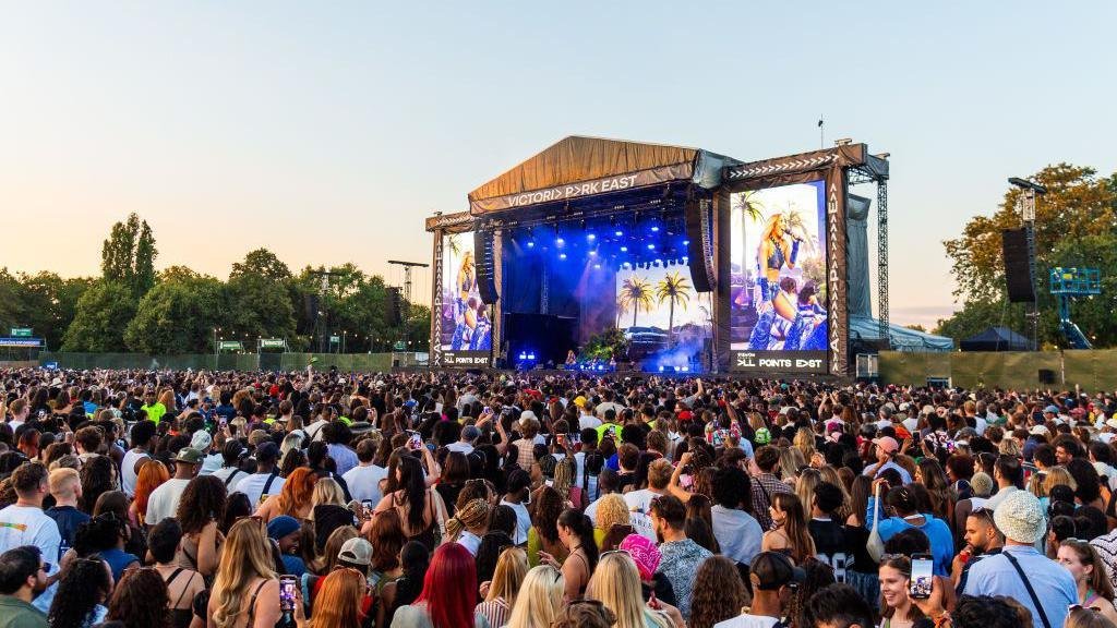 A large festival crowd watches a performer on the outdoor main stage at All Points East in Victoria Park under a clear evening sky.