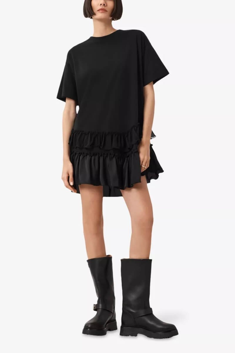 A model wearing AllSaints relaxed-fit black mini dress with frill hem and bold black knee-high boots