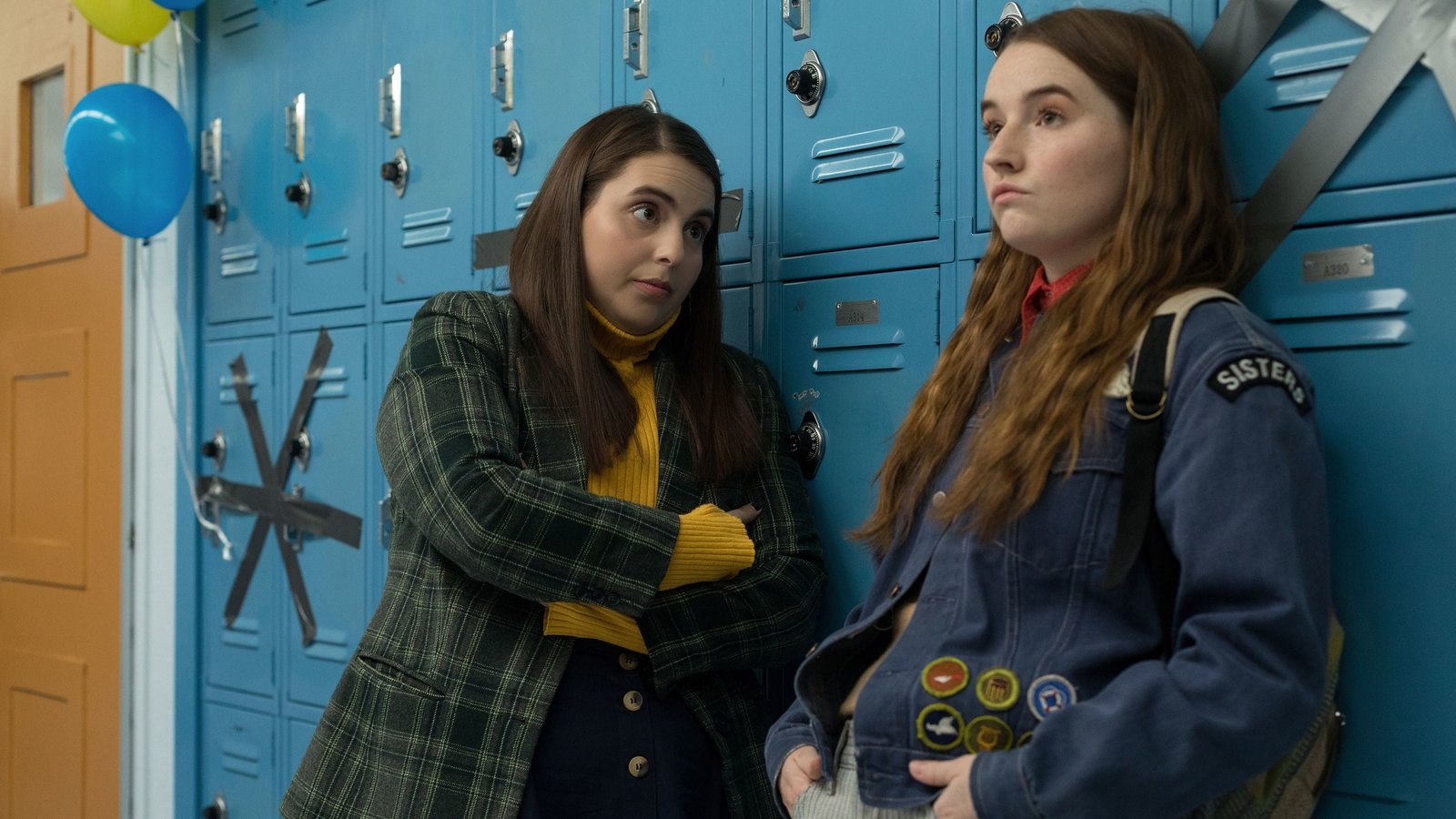 Beanie Feldstein and Kaitlyn Dever as Molly and Amy in a scene from 'Booksmart', leaning against bright blue high school lockers and sharing an expressive, tense moment.
