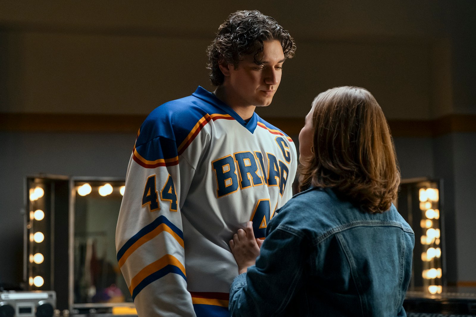 Belmont Cameli as Garrett, in a white 'Briar' hockey jersey, looks down thoughtfully at Ella Bright as Hannah, who wears a denim jacket and has her hand on his chest in a scene from 'Off Campus'.