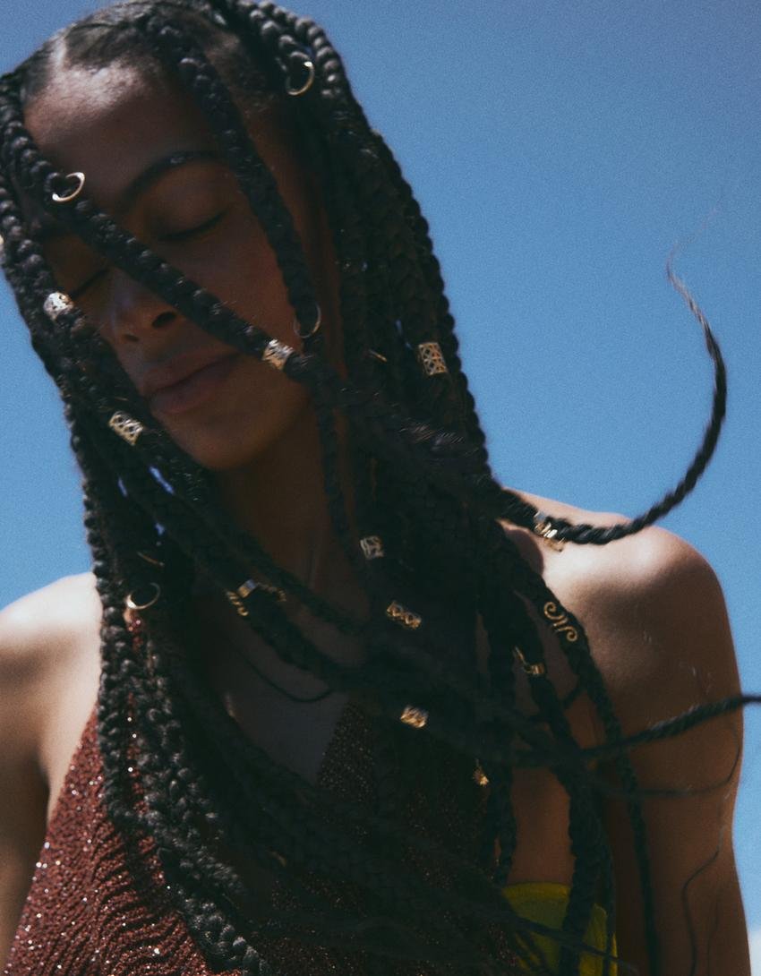 A woman with long braided hair adorned with accessories, looking down against a clear blue sky