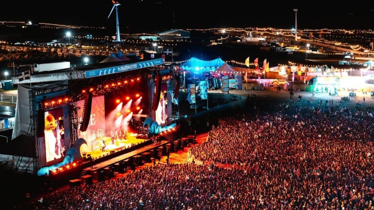 A high-angle nighttime view of a massive crowd packed in front of the brightly lit Main Stage at Boardmasters Festival, featuring wave-shaped stage decorations, large video screens, and a wind turbine visible in the illuminated background.