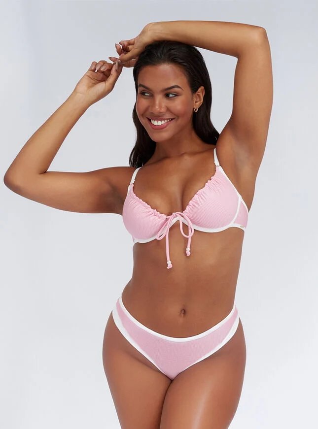 A smiling woman with dark, wavy hair is wearing the Boux Avenue Ibiza textured bikini in pink and white while posing with both arms raised against a plain white background in a studio photograph. The bikini top has gathered cups with white trim, a front tie detail, and underwire support for a bigger bust, while the high-leg bottoms feature a wide white waistband.