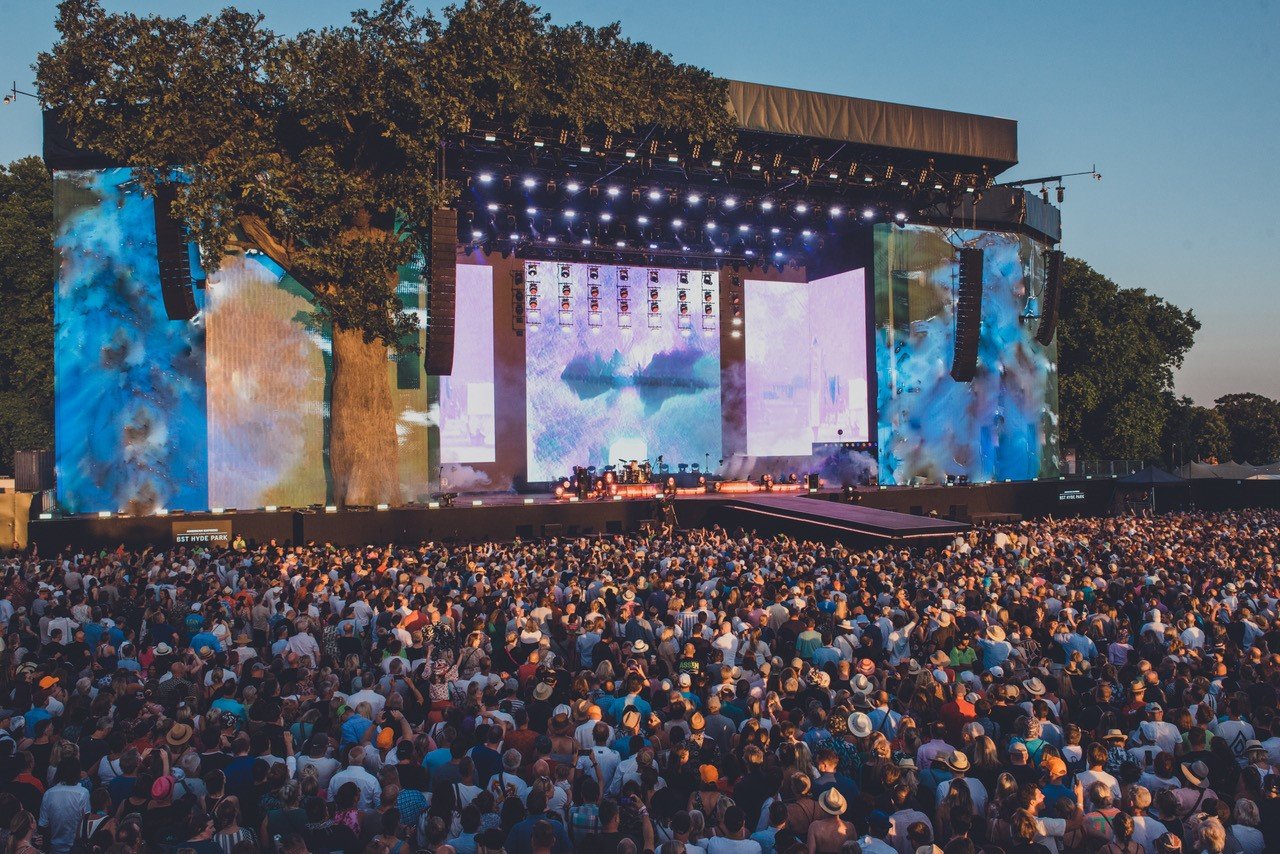 A massive crowd of people gathered in front of the Great Oak Stage at the BST Hyde Park music festival under a clear blue sky, with large video screens displaying blue and white visuals on either side of the stage.