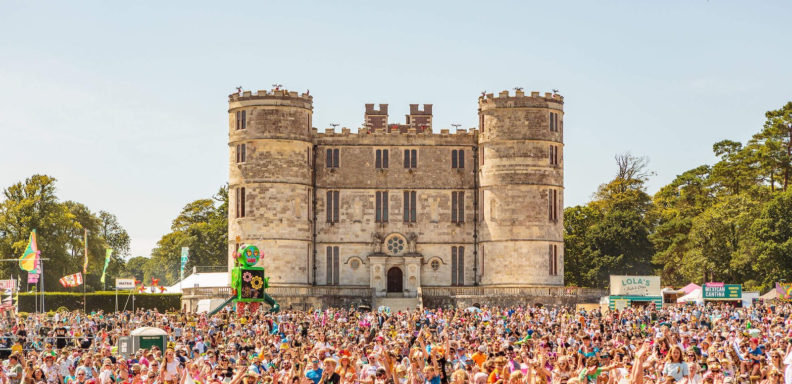 A massive crowd gathered on a sunny day in front of Lulworth Castle for Camp Bestival, surrounded by colorful flags and festival stalls.