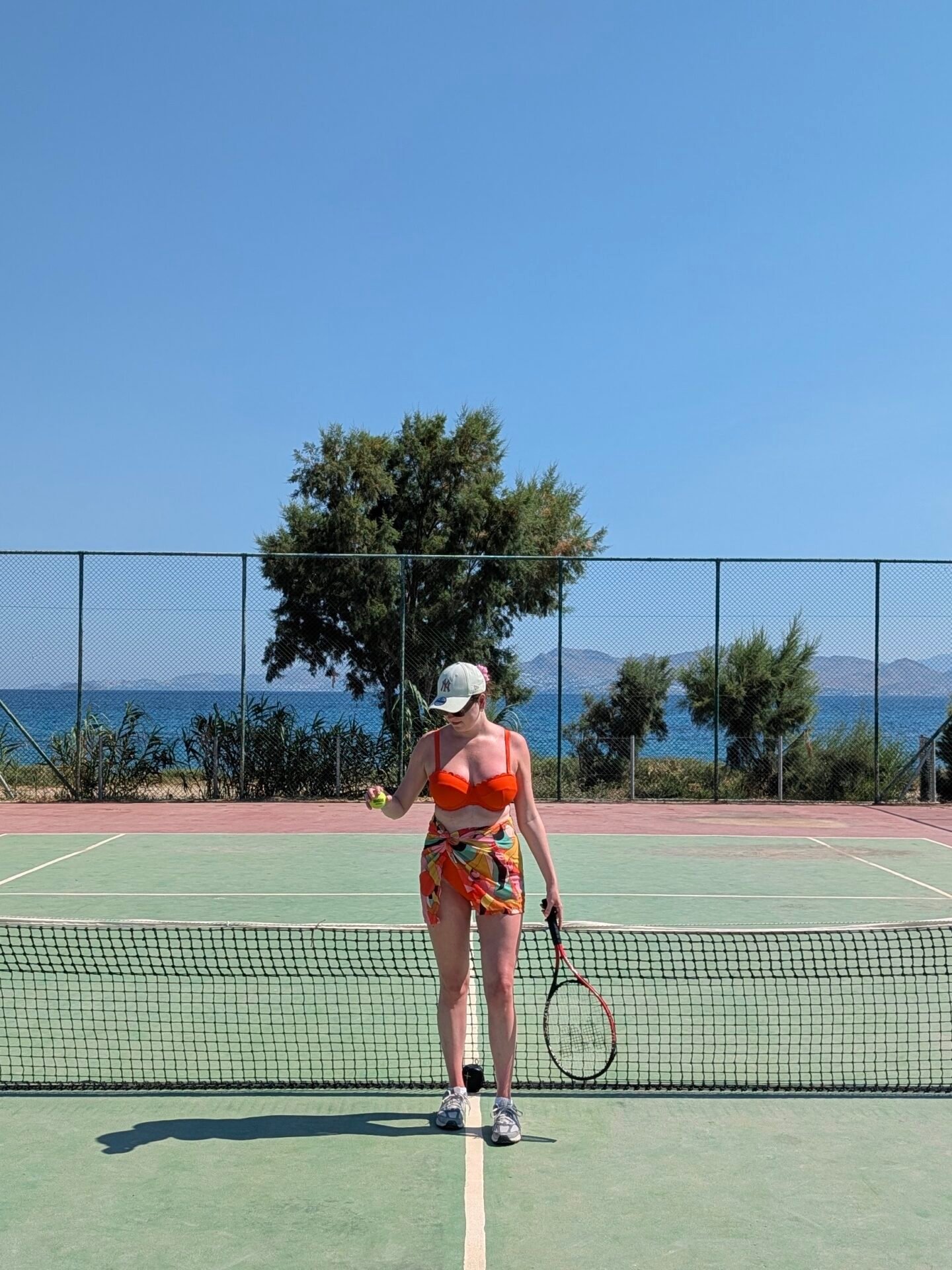 A woman stands at the net on an outdoor tennis court, wearing a bright orange JD Williams bikini top for DD+ boobs, a colourful patterned sarong, a baseball cap, and trainers. She is holding a tennis racket and a tennis ball, with a view of green trees and the blue sea behind the court's chain-link fence.
