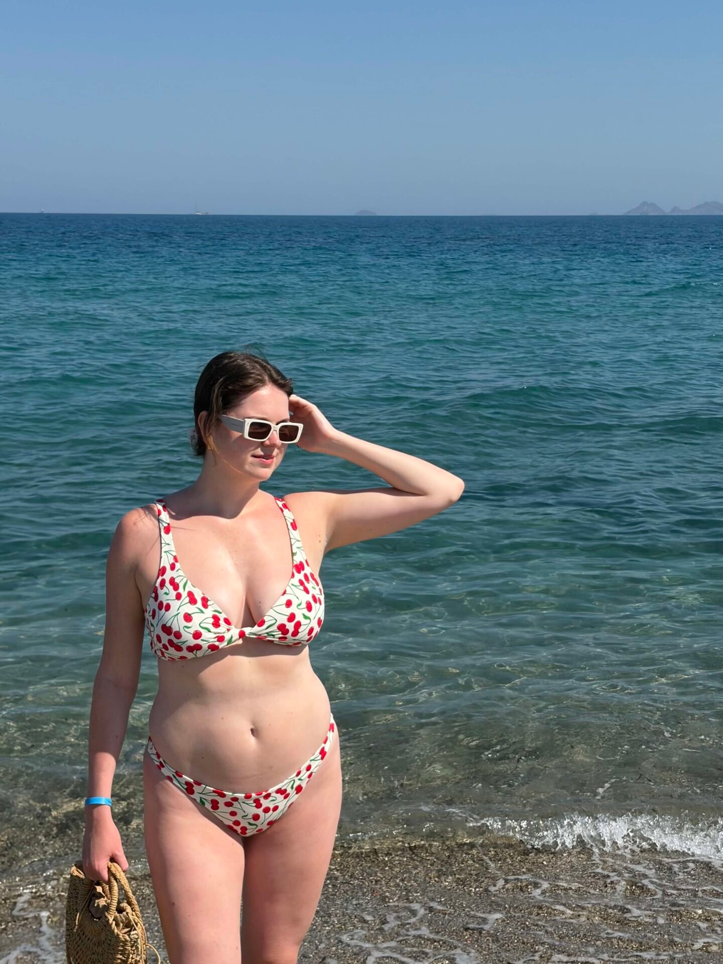 A woman stands on a sandy beach at the edge of the ocean, wearing white sunglasses and a cream-colored YITTY x Fabletics bikini DD+ with a red cherry print. She holds a woven straw bag in one hand while touching her hair with the other, with the blue sea and horizon in the background.