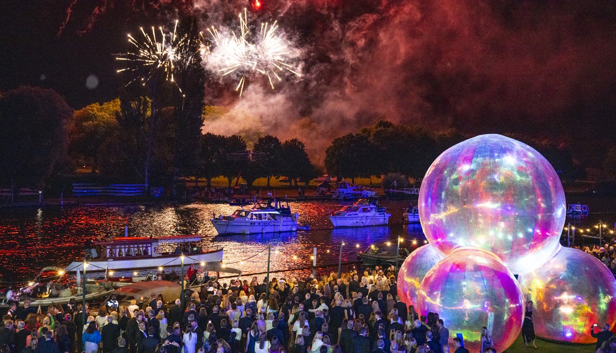 A formal-dressed crowd gathers along a riverbank at the Henley Festival at night to watch fireworks explode over boats on the water, with large, glowing iridescent spheres illuminating the foreground.