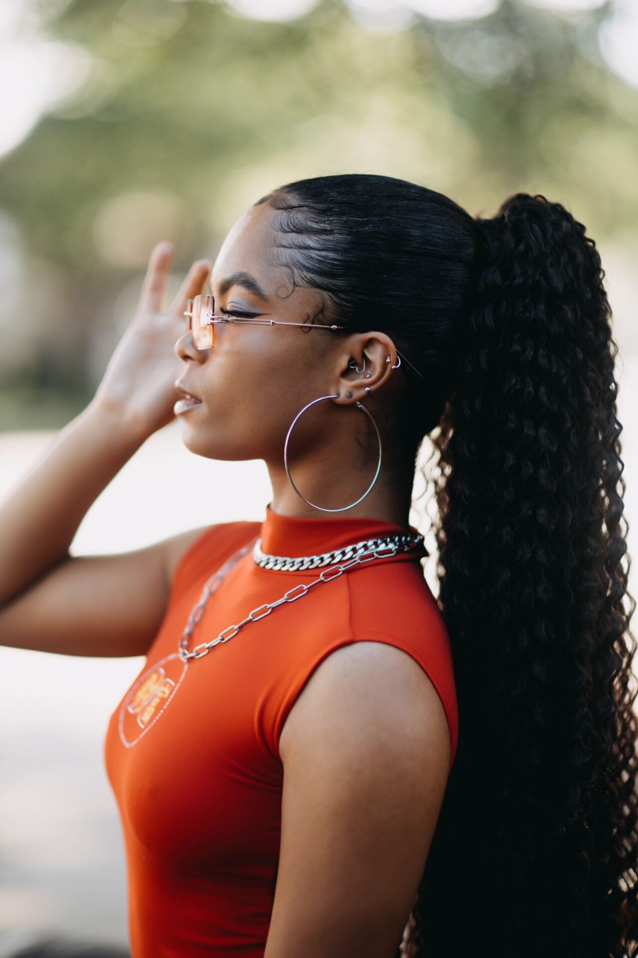 A young woman with long, curly hair styled in a high ponytail, wearing orange-tinted glasses and large hoop earrings, poses with her hand near her face, dressed in a fitted red top