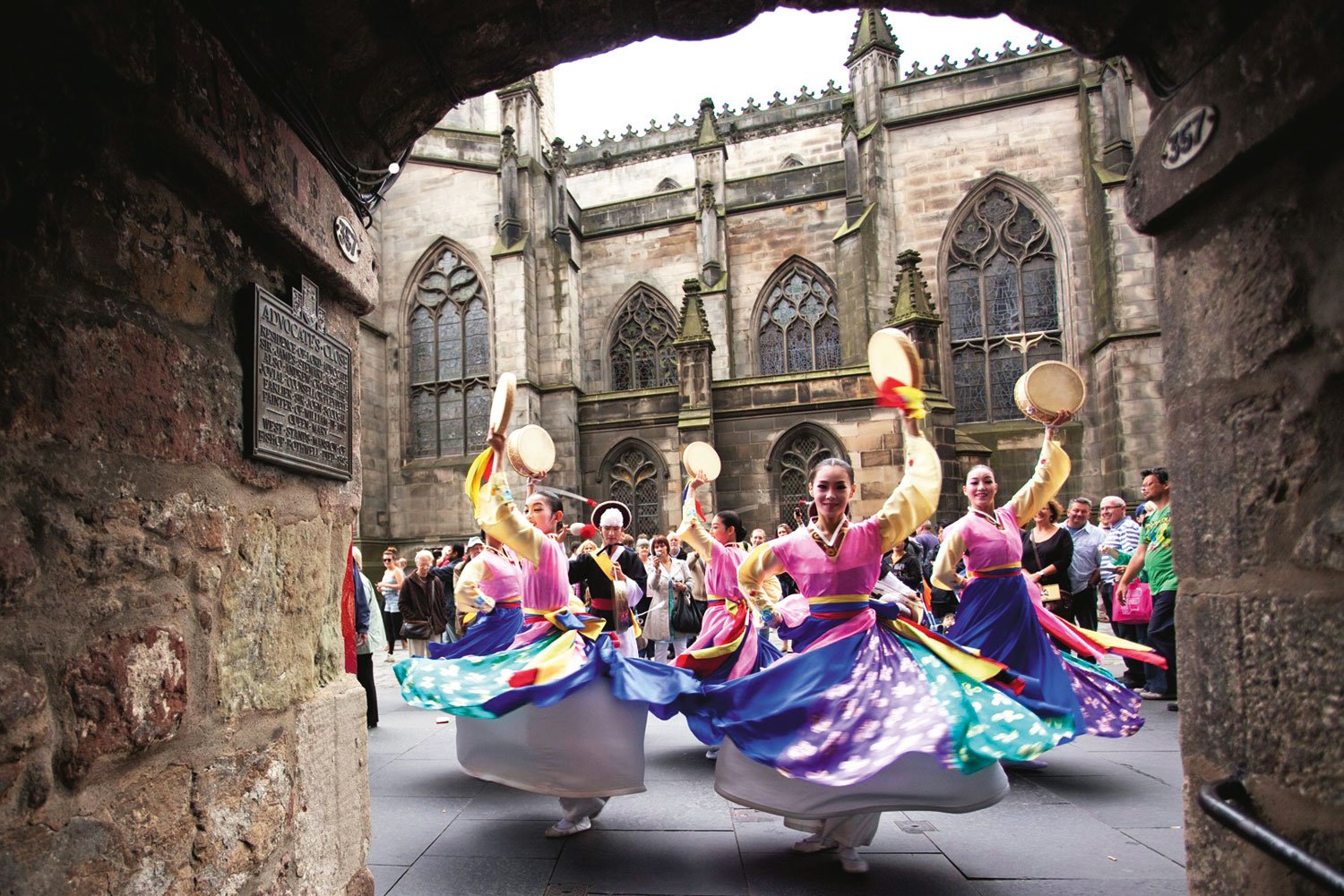 Traditional Korean dancers spin in vibrant dresses and hold drums while performing outdoors for a crowd in front of a historic stone building at the Edinburgh Festival Fringe.