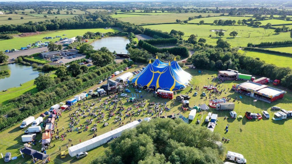 An aerial view of Deva Fest in Chester, featuring a large blue and yellow big top tent centered in a grassy field. The festival grounds include fairground attractions, vehicles, and scattered attendees, all surrounded by lush green countryside and a small lake.
