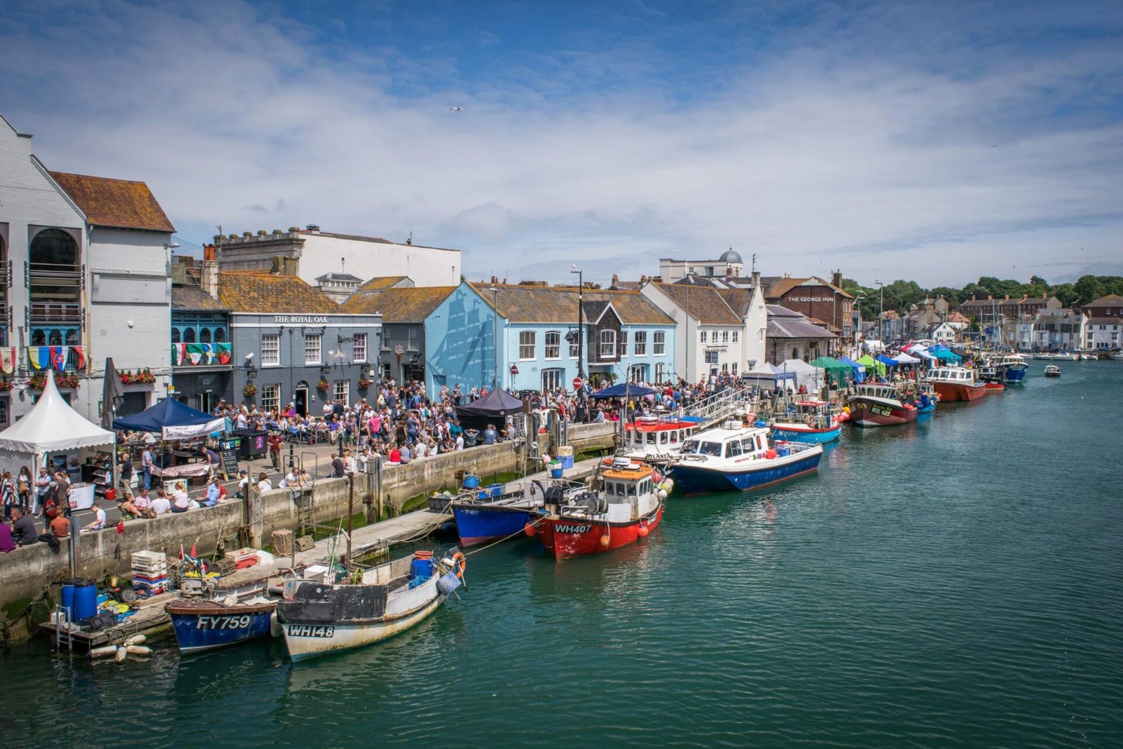 Crowds at the Dorset Seafood Festival along the Weymouth Harbour, with fishing boats moored by the quayside in front of colorful buildings.