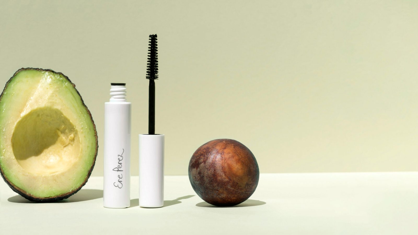 An open avocado half, a tube of Ere Perez mascara, and a brown avocado stone on a light green background - Ere Perez Avocado Waterproof Mascara beside fresh avocado