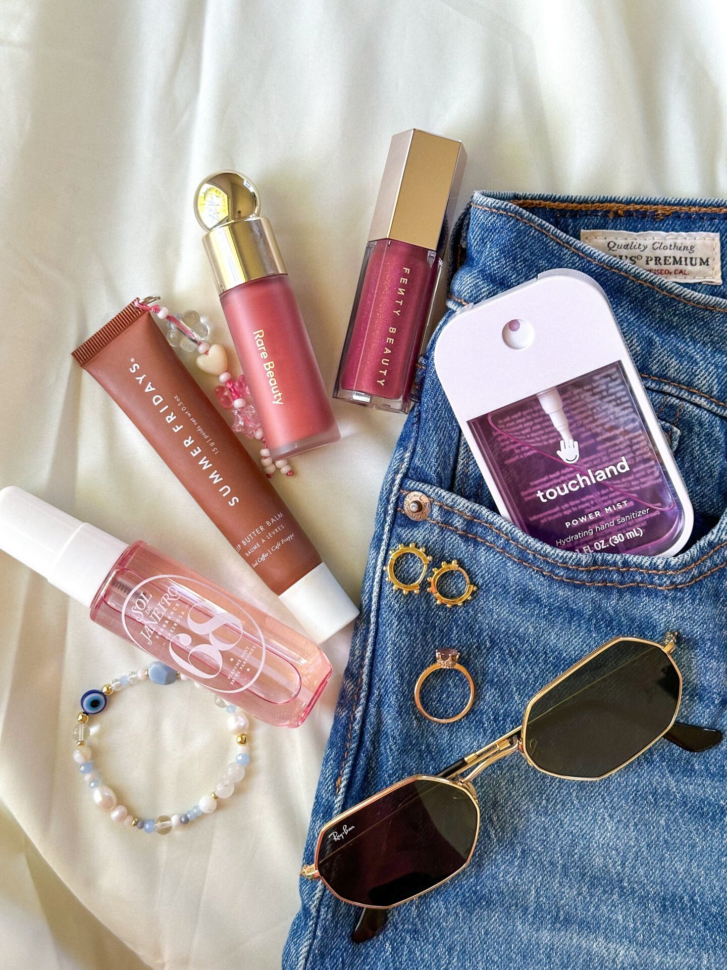 Array of beauty products and accessories on denim jeans. Includes lip glosses, hand sanitiser, sunglasses, delicate bracelet, rings, and a hair clip - best festival beauty essentials 2026