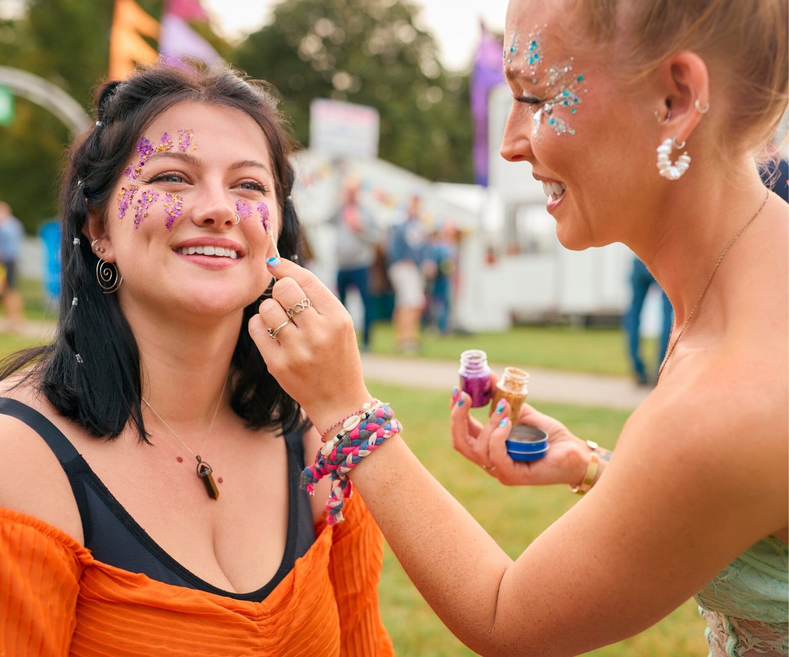 A smiling woman wearing an orange top has purple glitter and face gems applied to her cheeks by a festival makeup artist with blue glitter makeup holds pots of glitter at an outdoor summer festival - festival makeup looks