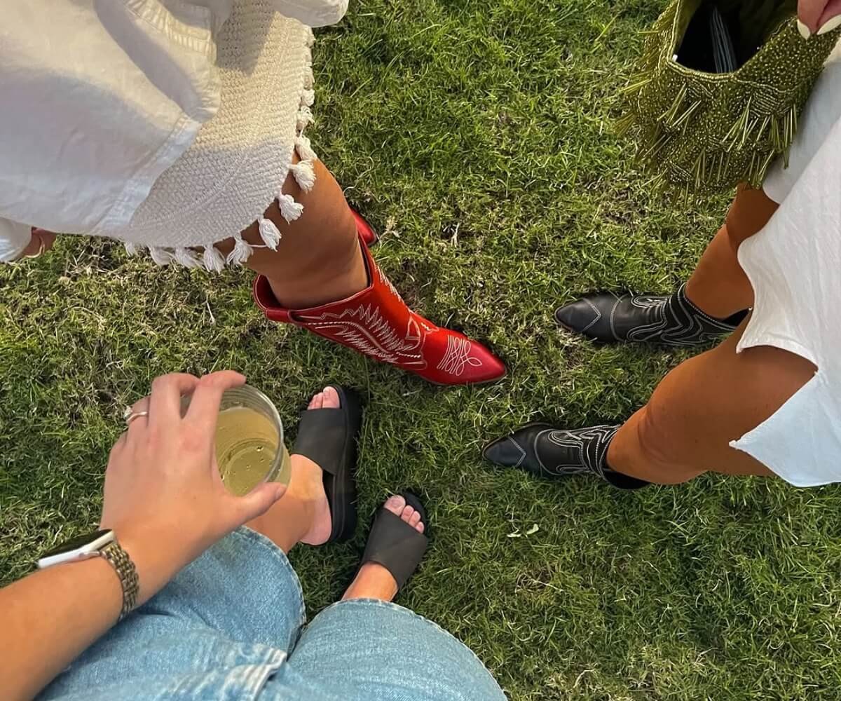 A group of three women standing on grass at a summer music festival wearing festival outfits, including red and black cowboy boots, boho-style dresses and casual denim. The image showcases popular festival fashion outfit ideas and trends for summer 2026, combining Western-inspired accessories with relaxed festival clothing