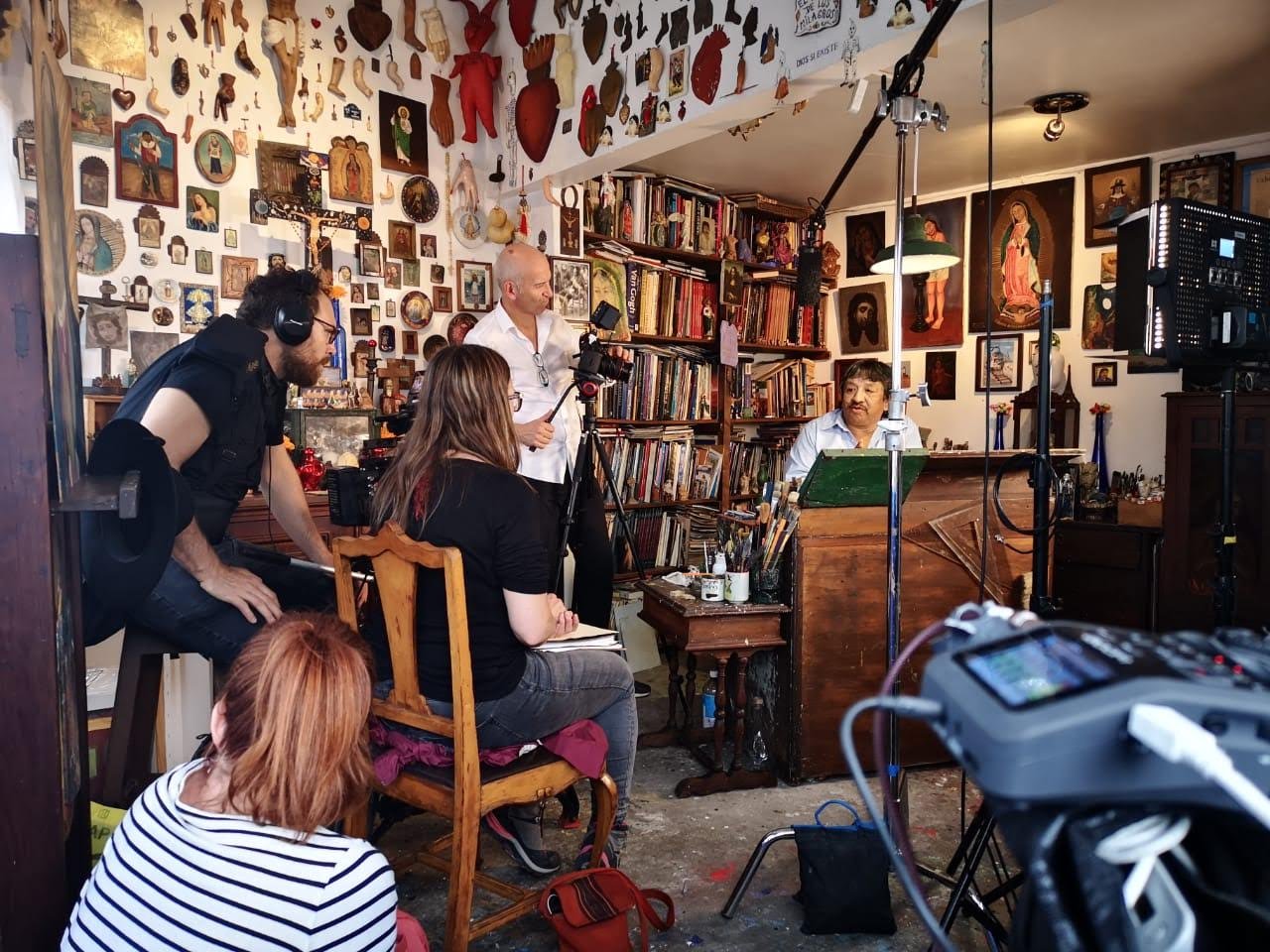 A behind-the-scenes view of a film crew interviewing Alfredo Vilchis Roque in a studio packed with books, religious icons, and anatomical wall art.