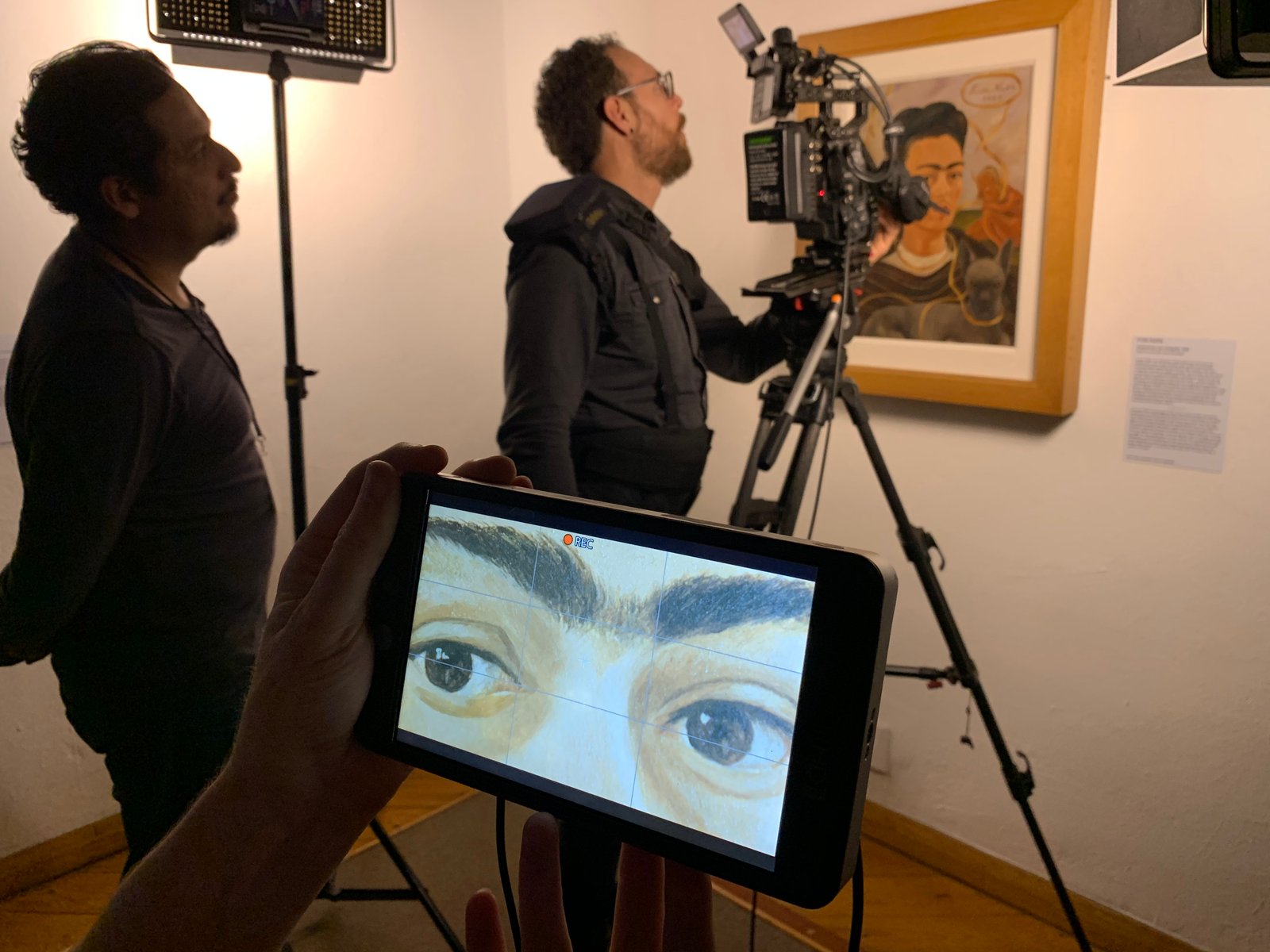 A behind-the-scenes view of a film crew filming a Frida Kahlo self-portrait in a gallery, with a handheld monitor in the foreground showing a close-up of the painted eyes.