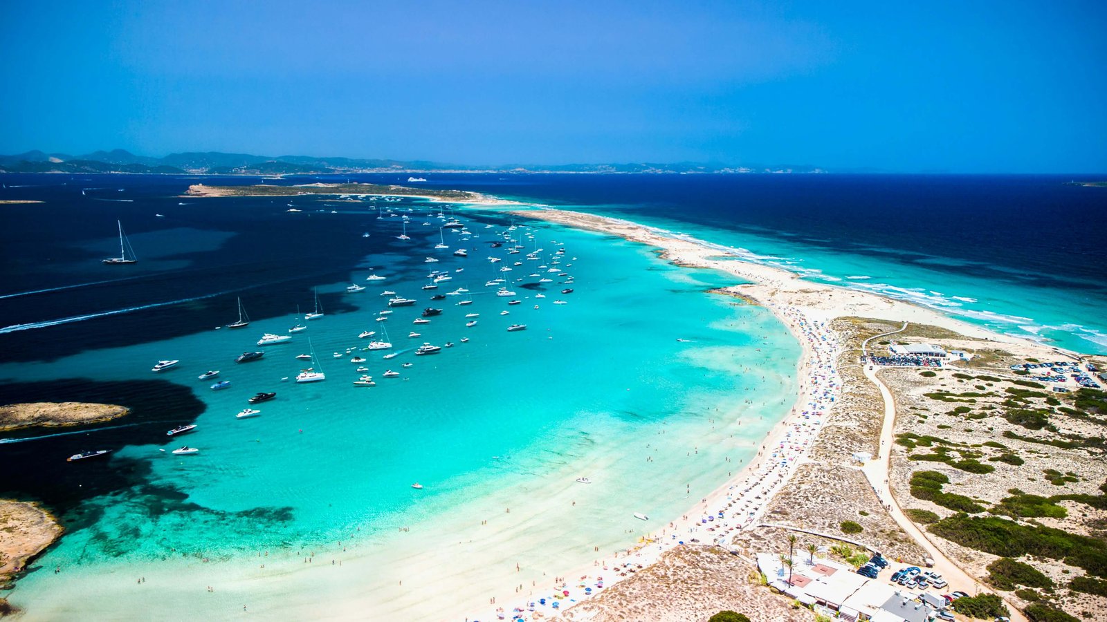 Aerial view of Ses Illetes beach in Formentera, Spain, with a long stretch of white sand surrounded by crystal-clear turquoise water, numerous boats anchored offshore, and people relaxing along the shoreline.