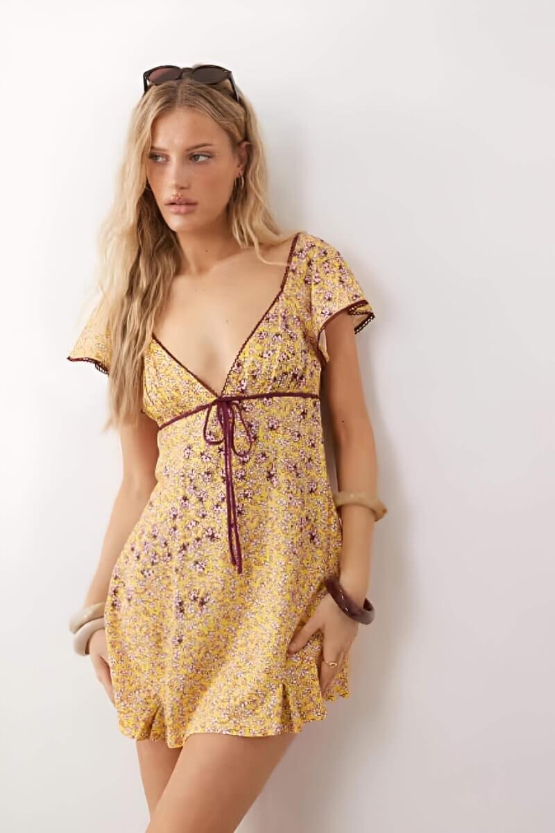 Model wearing Free People backroads flippy mini dress in dandelion, a yellow floral mini dress with a tied waist