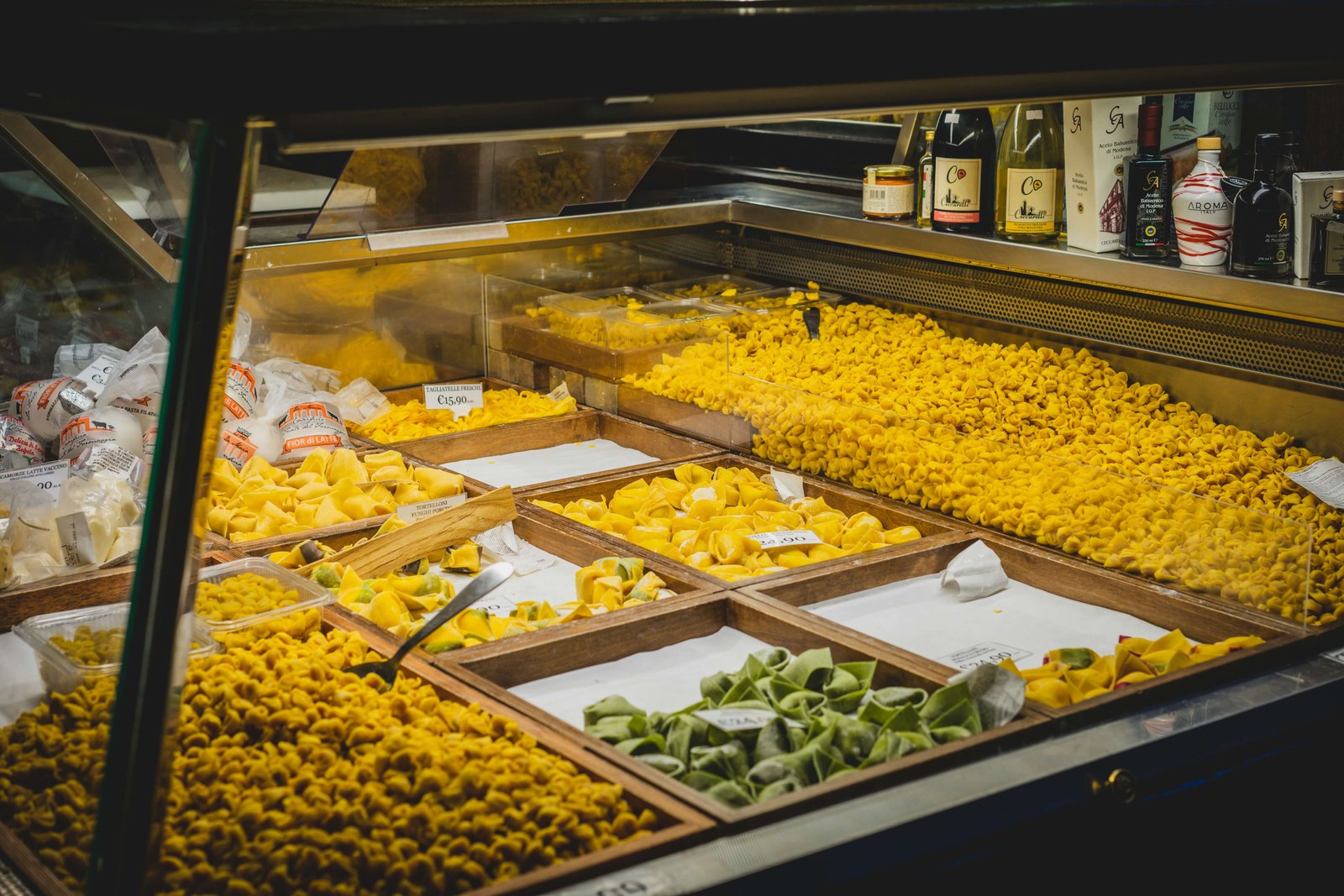 Assortment of fresh Italian pasta varieties displayed in a market counter, including tortellini, ravioli, and stuffed pasta in different colours, arranged in wooden trays under warm lighting.