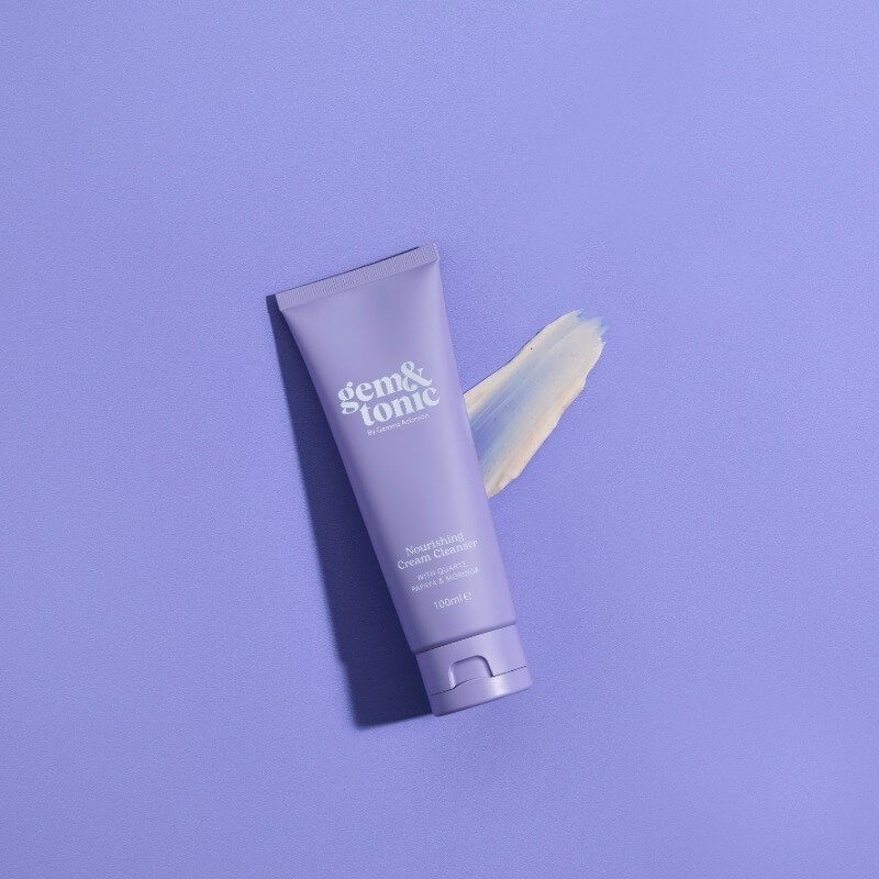 A purple tube of Gem & Tonic Nourishing Cream Cleanser with a cream swatch beside it on a matching background
