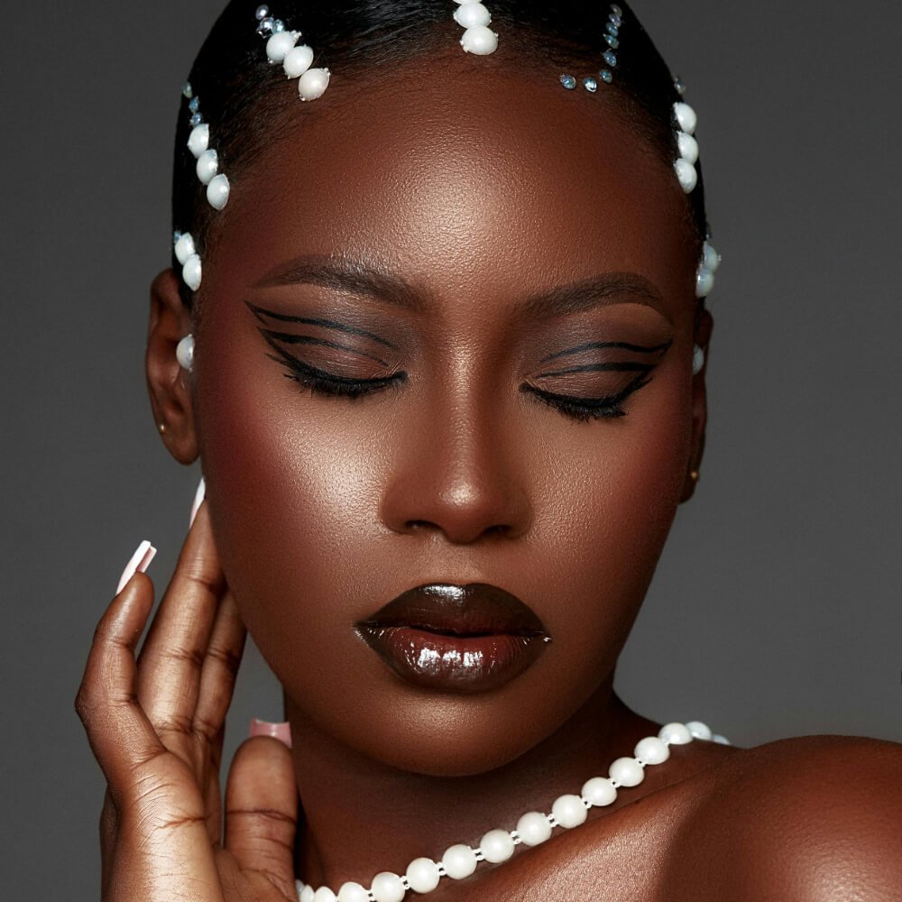 A woman with glossy dark berry lips, bold eyeliner, and a pearl-adorned hairstyle, showcasing a festival makeup look