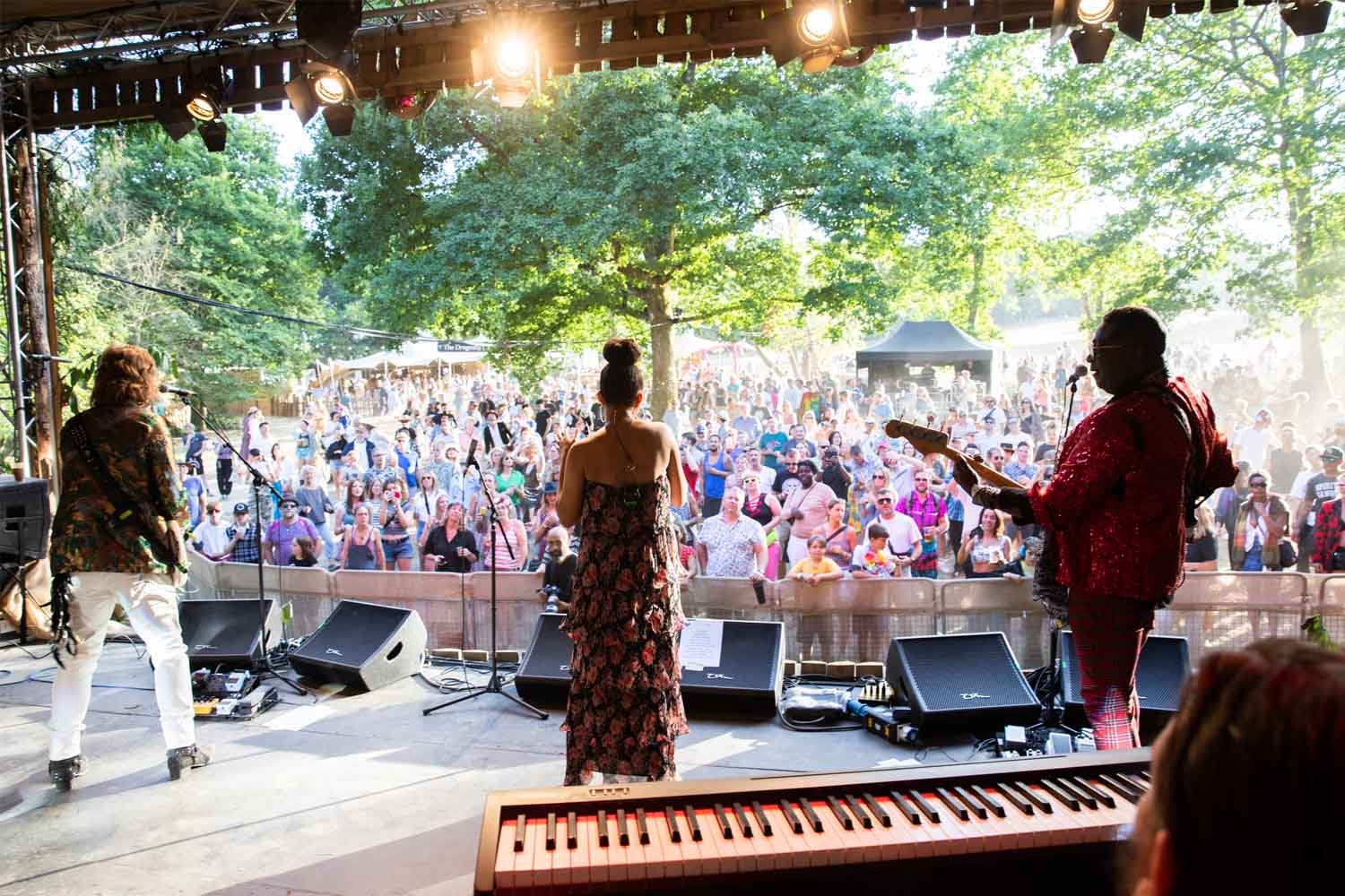 A band performs live on a shaded outdoor stage facing a large, sunlit crowd gathered in a wooded area at the Good Vibrations Society festival.