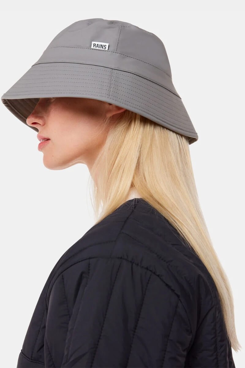 Model wearing Rains grey bucket hat, looking to the side, with blonde hair and a black jacket