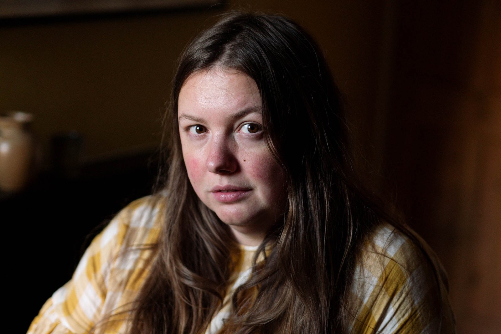 Portrait of Skins actress Hannah Murray now with a serious expression, featuring long brown hair and a yellow and white plaid top against a dark background.