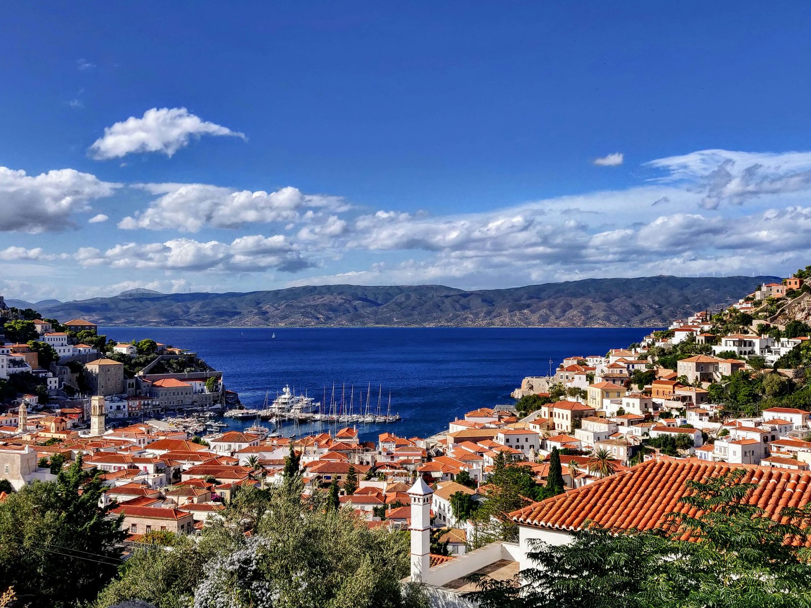 Panoramic view of Hydra Island in Greece showing white houses with red-tiled roofs cascading down hills toward a blue harbour filled with sailboats, with mountains visible across the sea under a bright sky.