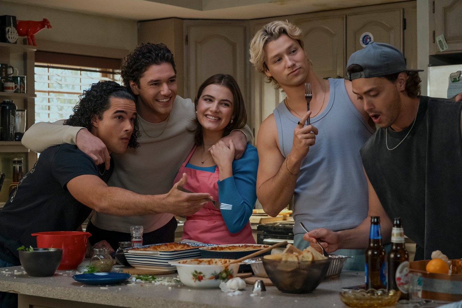 Jalen Thomas Brooks as Tucker, Belmont Cameli as Garrett, Ella Bright as Hannah, Stephen Kalyn as Dean, and Antonio Cipriano as Logan gathered in a cozy kitchen in a scene from 'Off Campus'. They are smiling and interacting around a counter filled with freshly prepared food, including casseroles and bread.