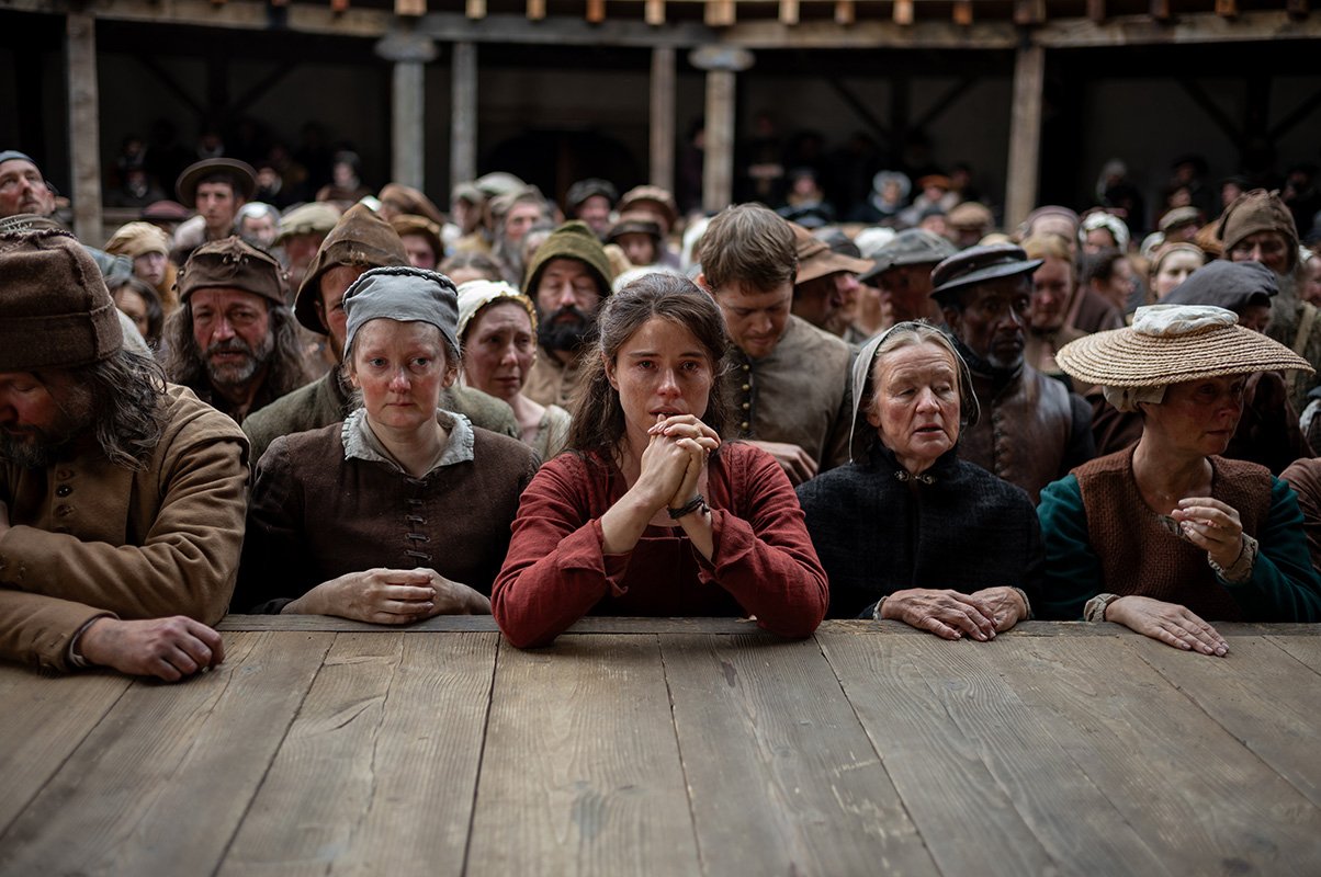 Jessie Buckley as Agnes is surrounded by a crowd, with her hands together in a prayer position, leaning onto the stage to watch a play in a scene from 'Hamnet'.