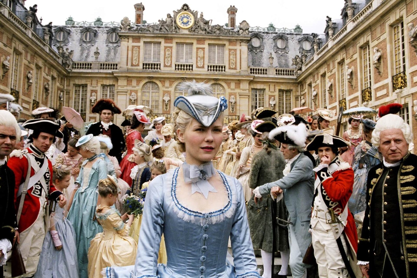 Kirsten Dunst as the titular queen in 'Marie Antoinette', standing in a crowded courtyard at the Palace of Versailles, wearing an ornate blue 18th-century gown and matching hat.