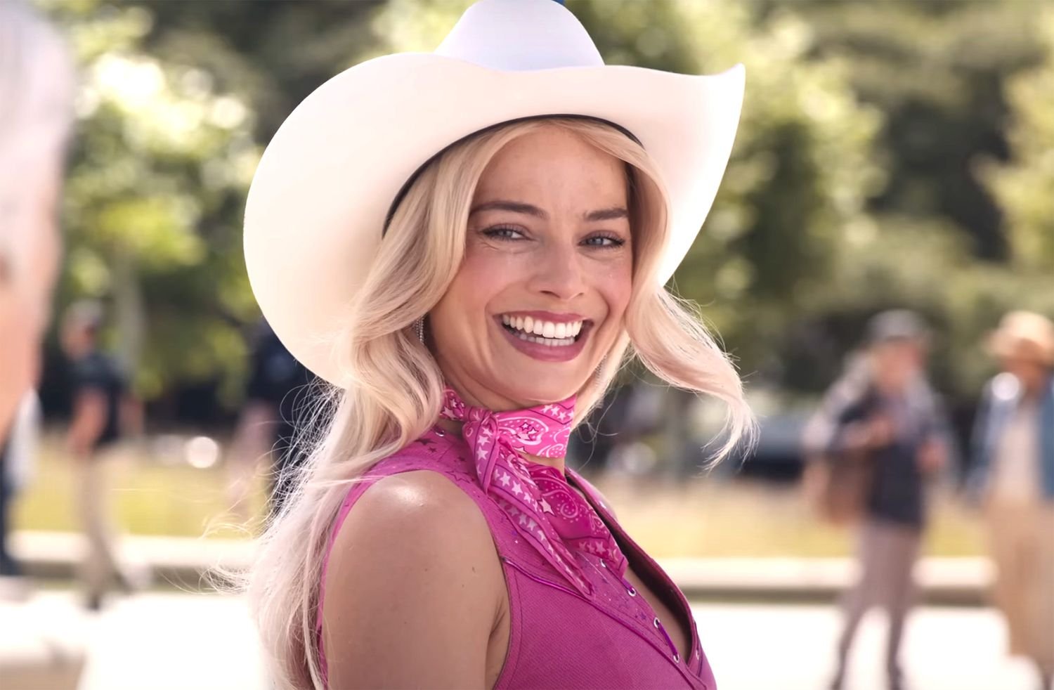 Margot Robbie as Barbie, wearing a white cowboy hat and a pink bandana around her neck, smiling in a scene from 'Barbie'.