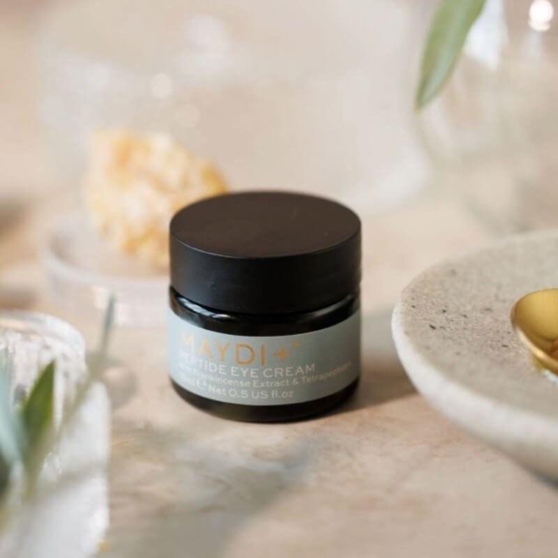 A small jar of MAYDI+ Peptide Eye Cream on a light stone surface with natural elements in the background