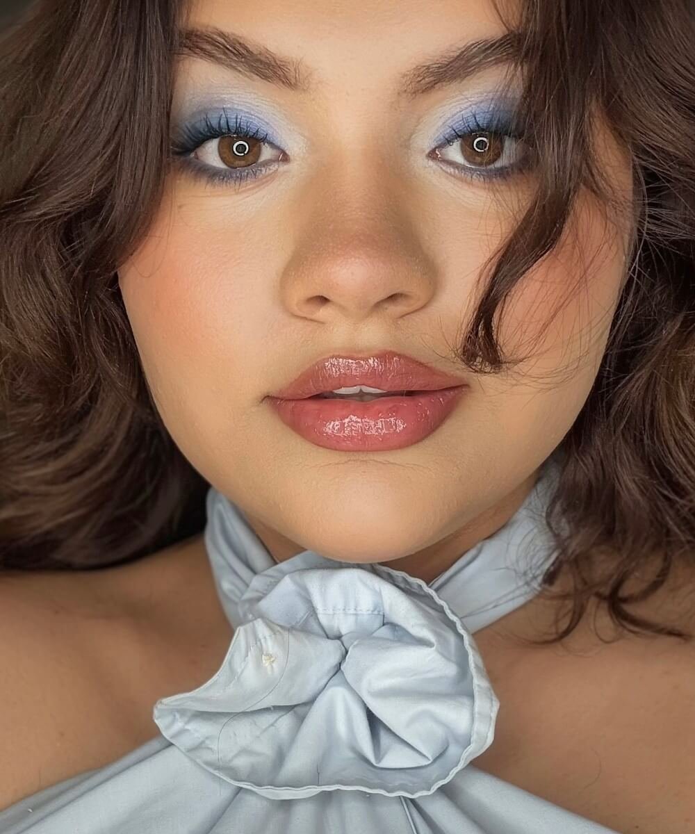 Close-up of a person with blue eyeshadow and glossy lips, wearing a light blue top with a bow