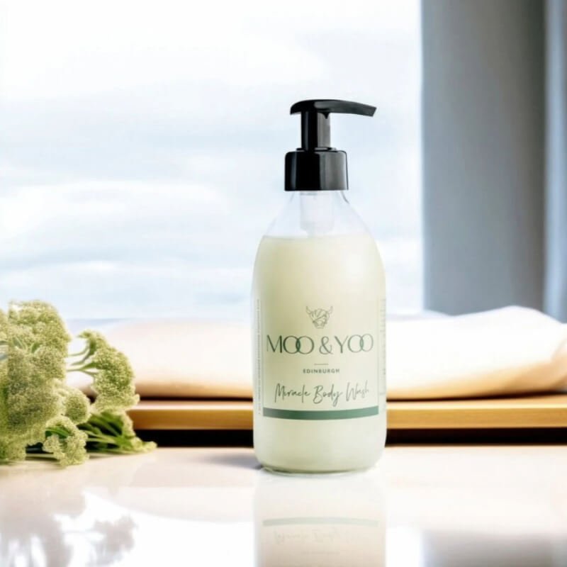 Bottle of Moo & Yoo Miracle Body Wash on a wooden surface beside decorative flowers, with light background